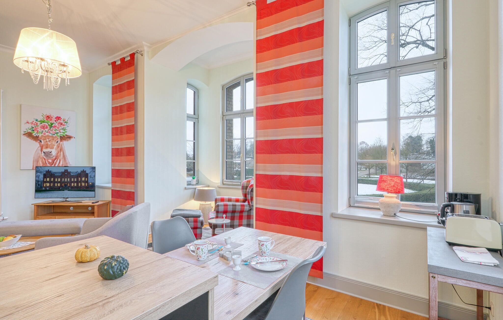Ein Essbereich mit einem Holztisch, gedeckt für vier Personen, einem Fernseher auf einem Holzschrank und einem Fenster mit Blick auf Bäume und ein Gebäude.