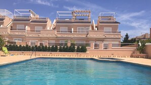 A swimming pool with a clear blue water surface, surrounded by a low stone wall. In the background, there are multi-story buildings with balconies and windows. - 3027 Vista Azul,close to the beach &  heated pool (Torre de la Horadada)