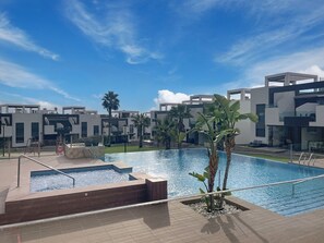 Un espace piscine moderne, avec un ciel d’un bleu limpide, entouré de bâtiments contemporains et de palmiers.