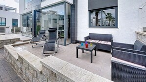 Un espace extérieur aménagé en terrasse, doté de mobilier moderne, notamment de fauteuils et d’un canapé, ainsi qu’un espace intérieur clos par des parois de verre, avec des tables et des chaises.