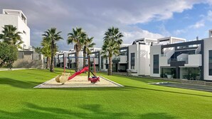 Un bâtiment moderne doté d’une aire de jeux avec un toboggan rouge et des balançoires, le tout entouré d’une pelouse bien entretenue et de palmiers.