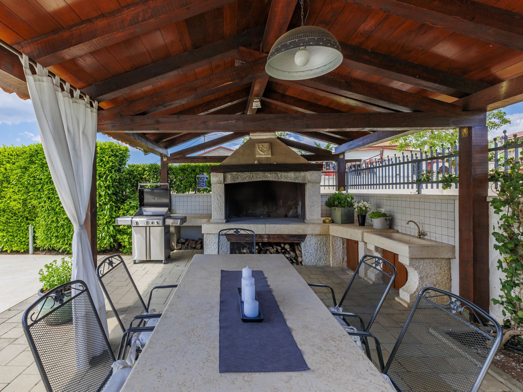 Un patio al aire libre con una parrilla de piedra, sillas de metal, una mesa con velas y un colgador de luces.