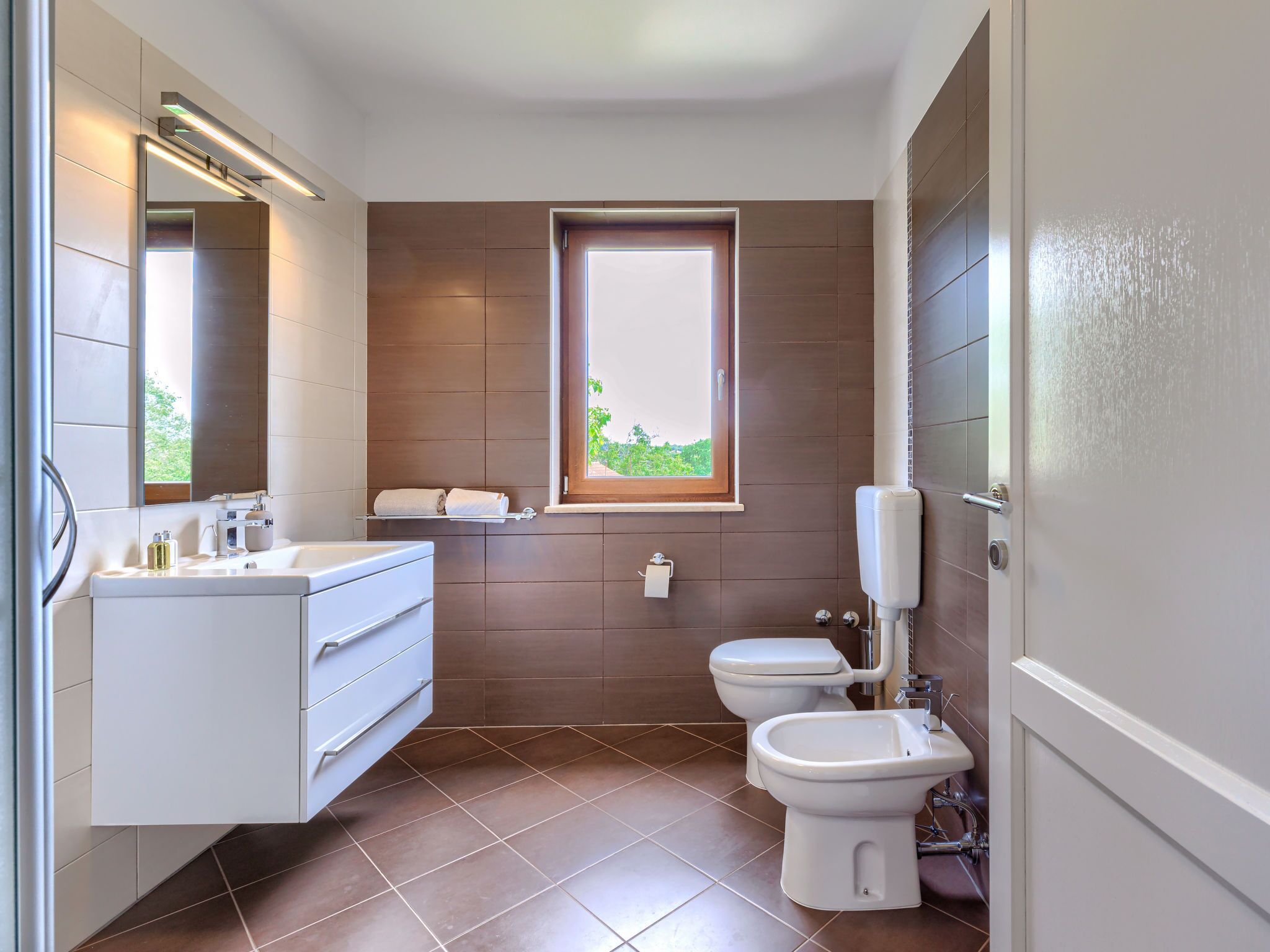 A modern bathroom with a white vanity, a toilet, and a bidet. There is a window providing natural light and a view of greenery outside.