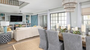 Ein geräumiges Wohnzimmer mit großem Fernseher, grauen Polsterstühlen, einem Holzessttisch mit Kerzen und einer Kristallleuchter.