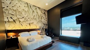 Una habitación de hotel moderna con una cama grande, un panel decorativo, una mesita de noche, una silla y un ventanal con vistas a la ciudad.