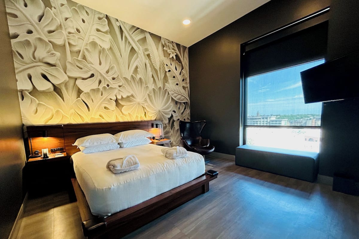 A modern hotel room with a large bed, a decorative wall, a nightstand, a chair, and a large window with a city view.