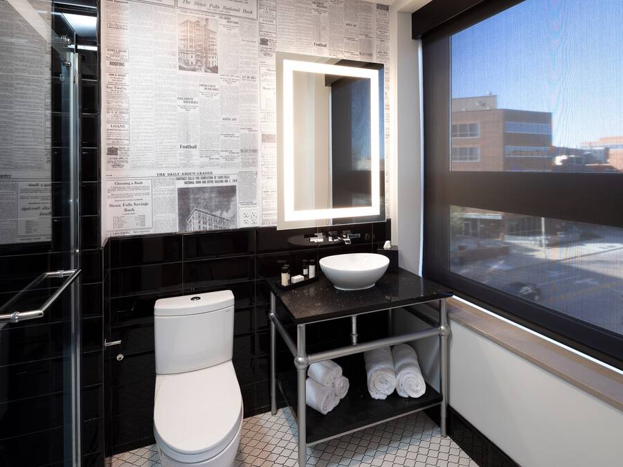 Un baño moderno con pared de azulejos negros, un inodoro blanco, un lavamanos con espejo y una ventana con vistas a edificios.