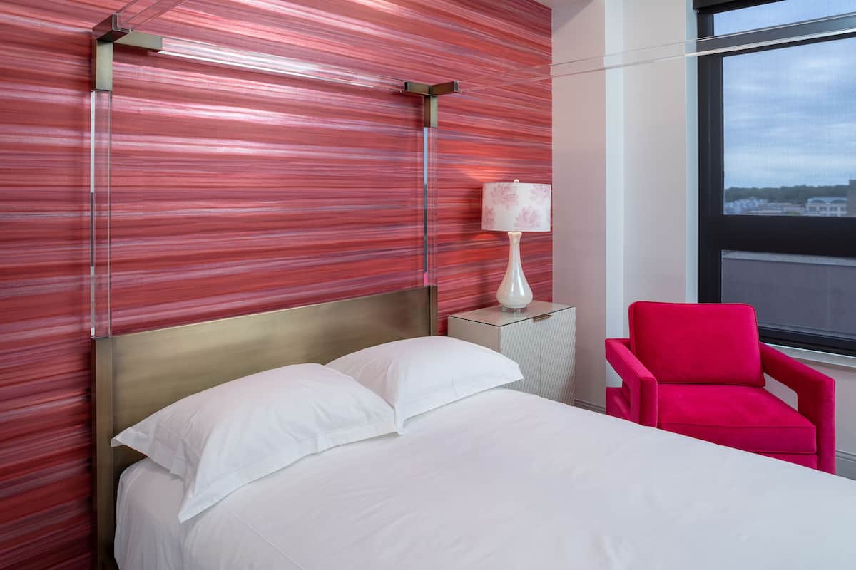 A hotel room with a bed, a red chair, a nightstand with a lamp, and a window with a view of buildings.