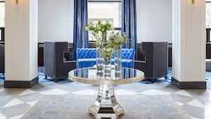 Un vestíbulo con una gran lámpara de araña, cortinas azules y una mesa redonda de vidrio con un jarrón de flores.