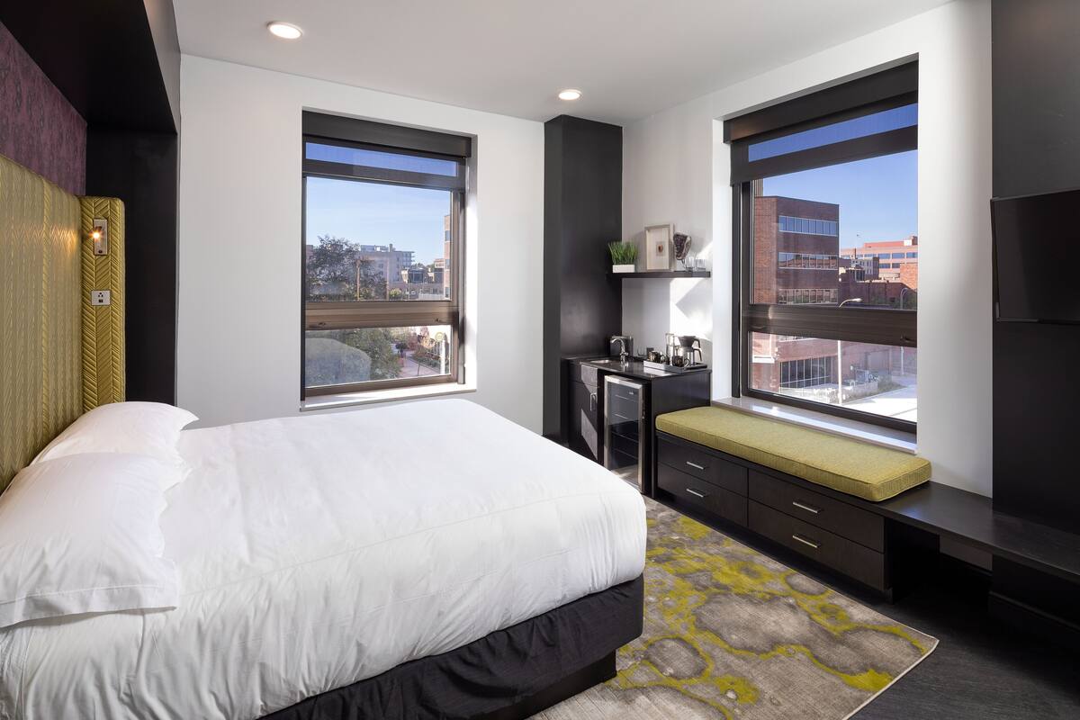 A modern bedroom with a large bed, a desk with a bench, and a view of the city through large windows.