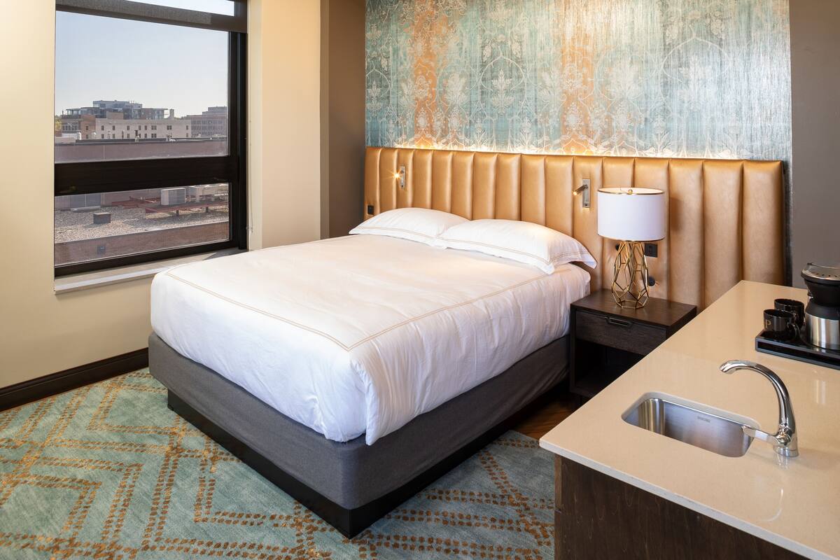 A hotel room with a bed, a nightstand, a lamp, a sink, and a view of the city through a window.
