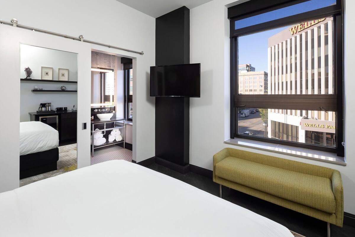 A hotel room with a bed, a TV, a bench, a window with a view of Wells Fargo building, and a bathroom with a sink and mirror.