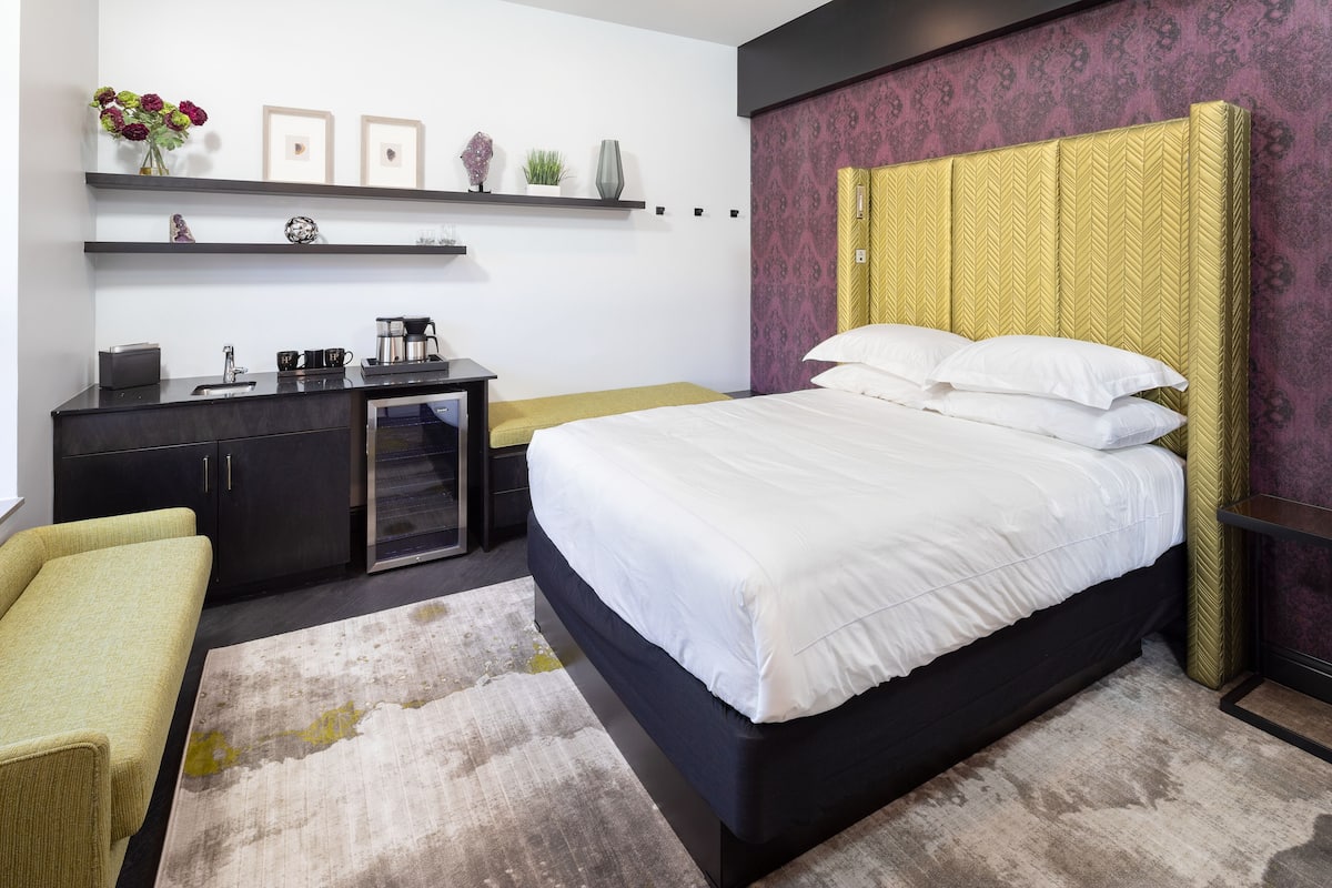 A modern bedroom with a bed, bedside table, and a desk with a chair. There are shelves above the bed and a small table with a plant.