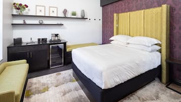 A modern bedroom with a bed, bedside table, and a desk with a chair. There are shelves above the bed and a small table with a plant.