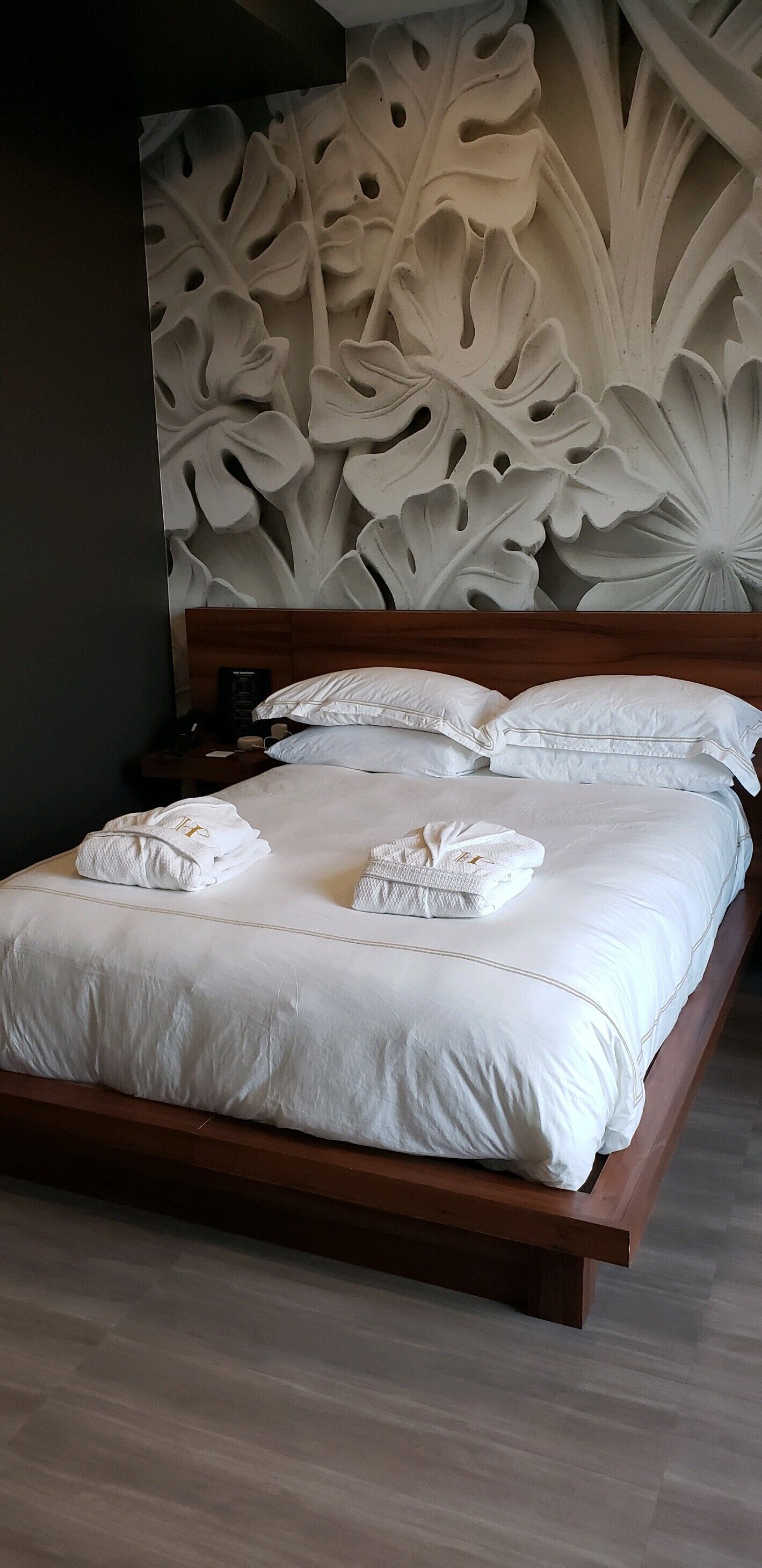 A neatly made bed with white linens and towels, a wooden headboard, and a wall with a large floral carving.