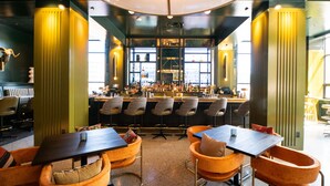Zona de bar con taburetes y mesas altas, barra con botellas y un área de estar con sillones acolchados.