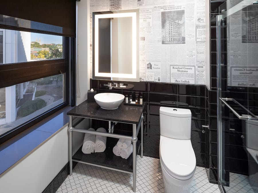 Un baño moderno con un espejo grande, un lavabo blanco y un inodoro. Una ventana ofrece vistas de edificios exteriores.