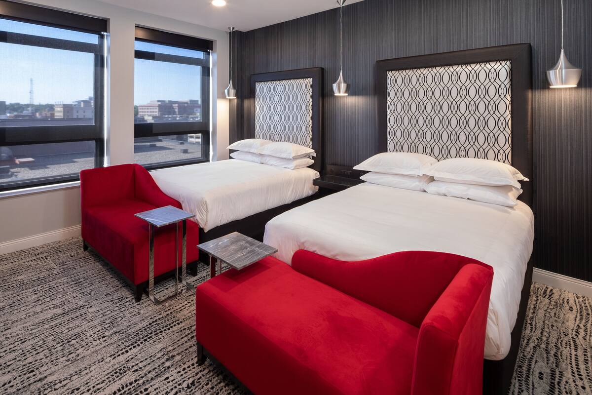 A hotel room with two beds, a red sofa, a small table, and large windows offering a city view.