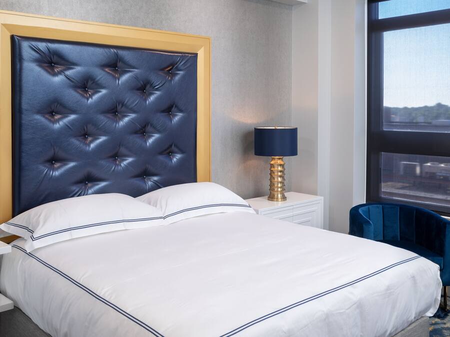 Et hotelværelse med en betrukken sengegavl, hvide sengetøj, et natbord med en lampe og udsigt gennem et vindue.