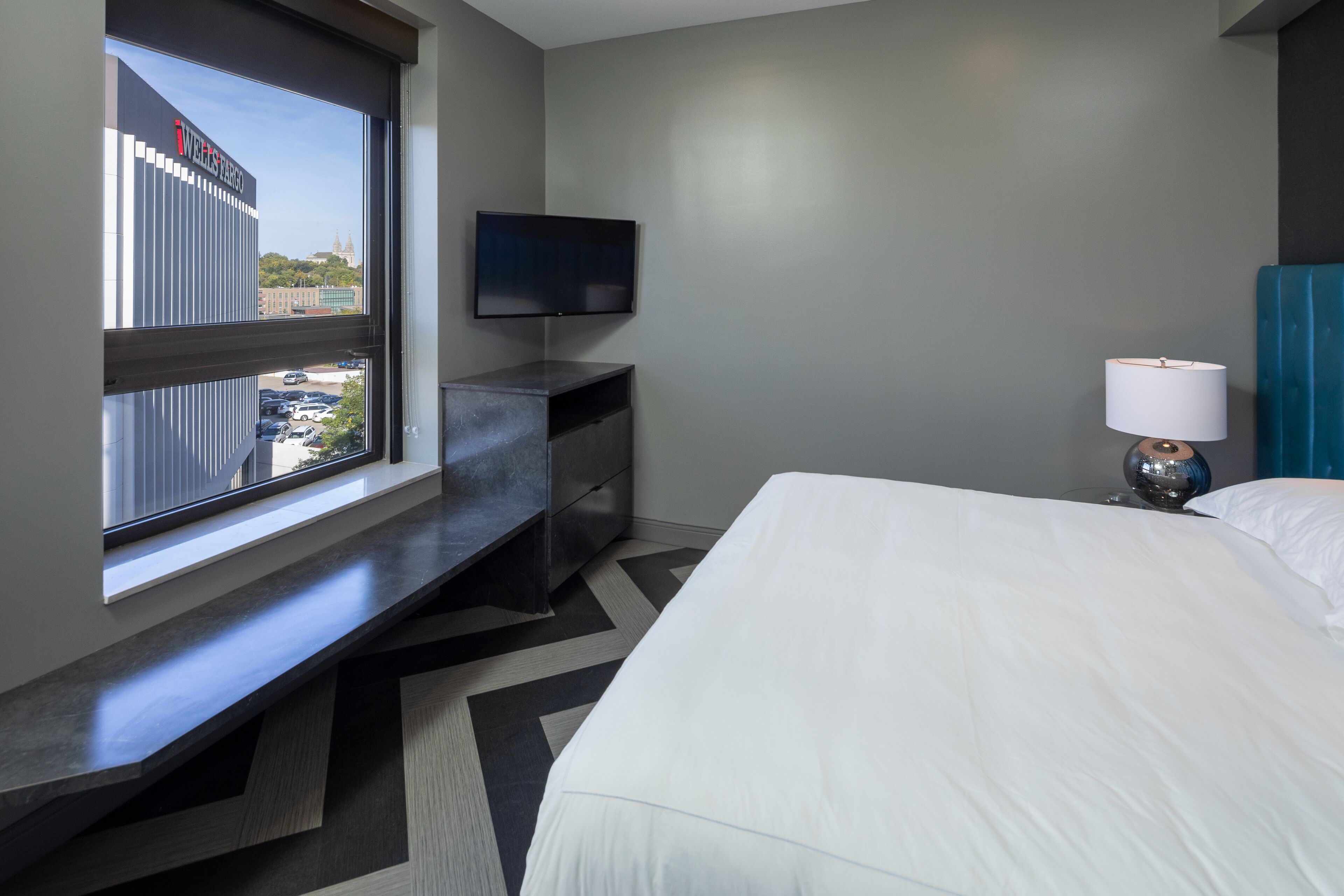 Quarto de hotel com cama, televisão na parede, secretária com cadeira e janela com vista para um edifício e carros estacionados.