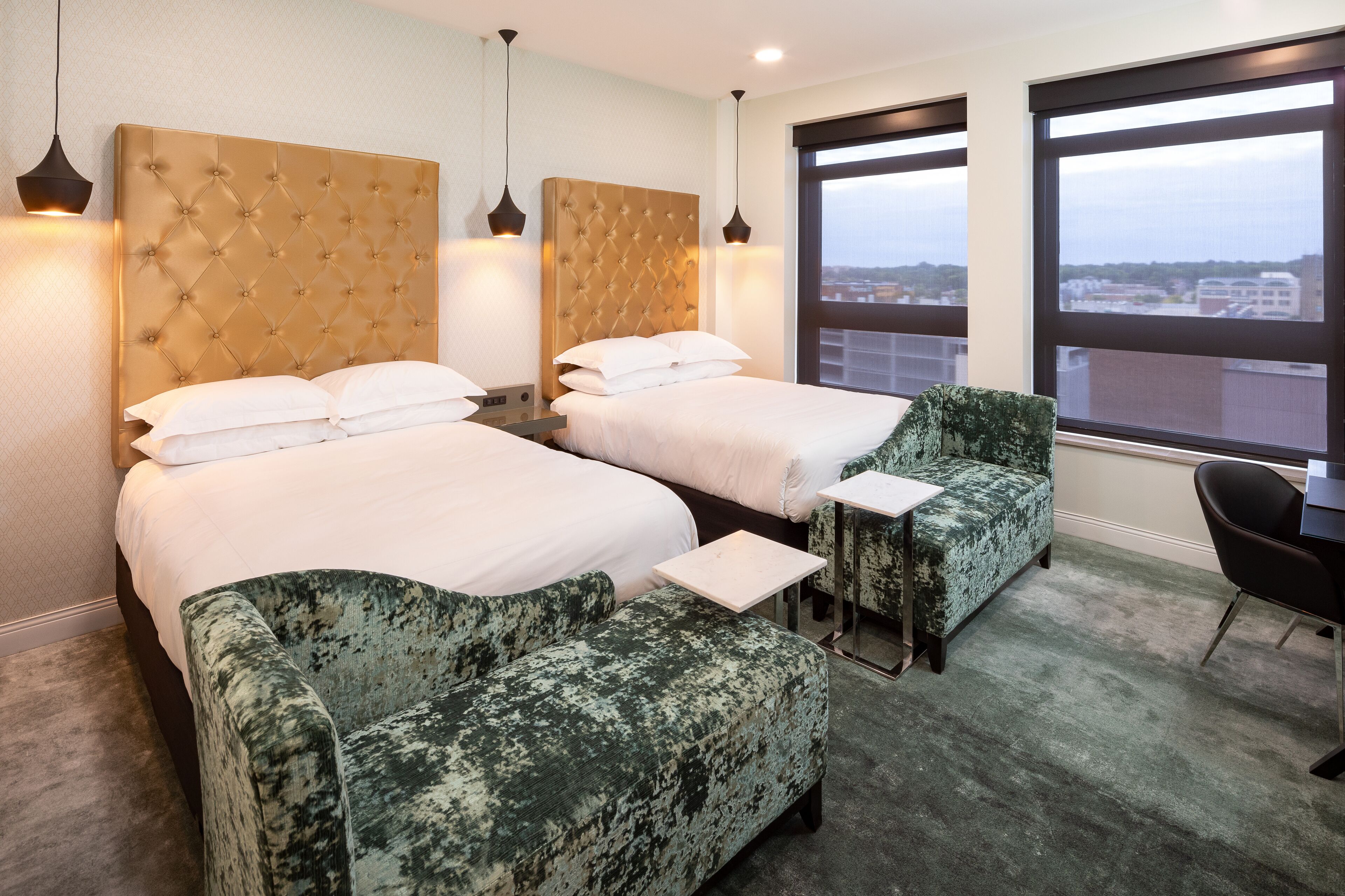 A hotel room with two beds, a green armchair, a small table, and a view of the city through large windows.
