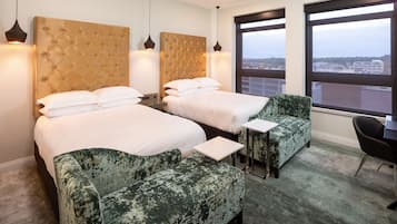 A hotel room with two beds, a green armchair, a small table, and a view of the city through large windows.