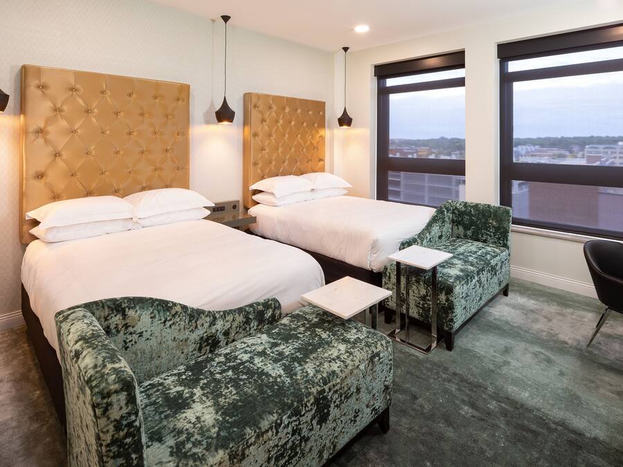 Une chambre d’hôtel avec deux lits, un fauteuil vert, une petite table et une vue sur la ville grâce à de grandes fenêtres.