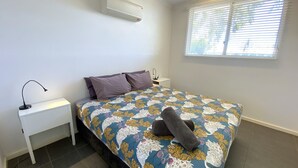 Een slaapkamer met een bed, nachtkastje, lamp en een raam met jaloezieën.