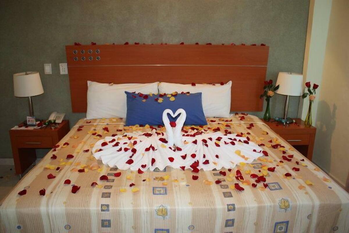 A hotel room with a bed covered in rose petals arranged in a heart shape, a blue pillow, and a wooden headboard.