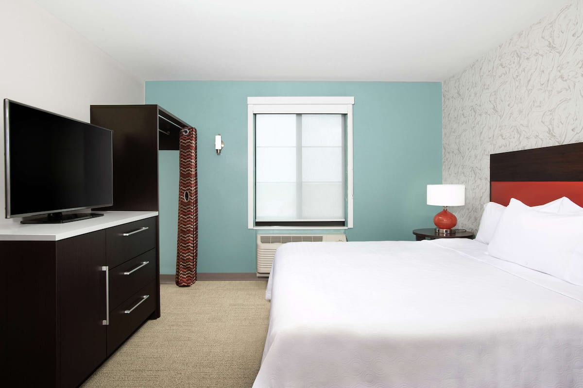 A hotel room with a bed, a TV on a dresser, a window, and a nightstand with a lamp.