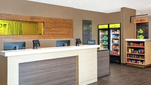 A hotel reception area with a modern design, including a reception desk, a digital payment terminal, a minibar, and a self-service beverage station.
