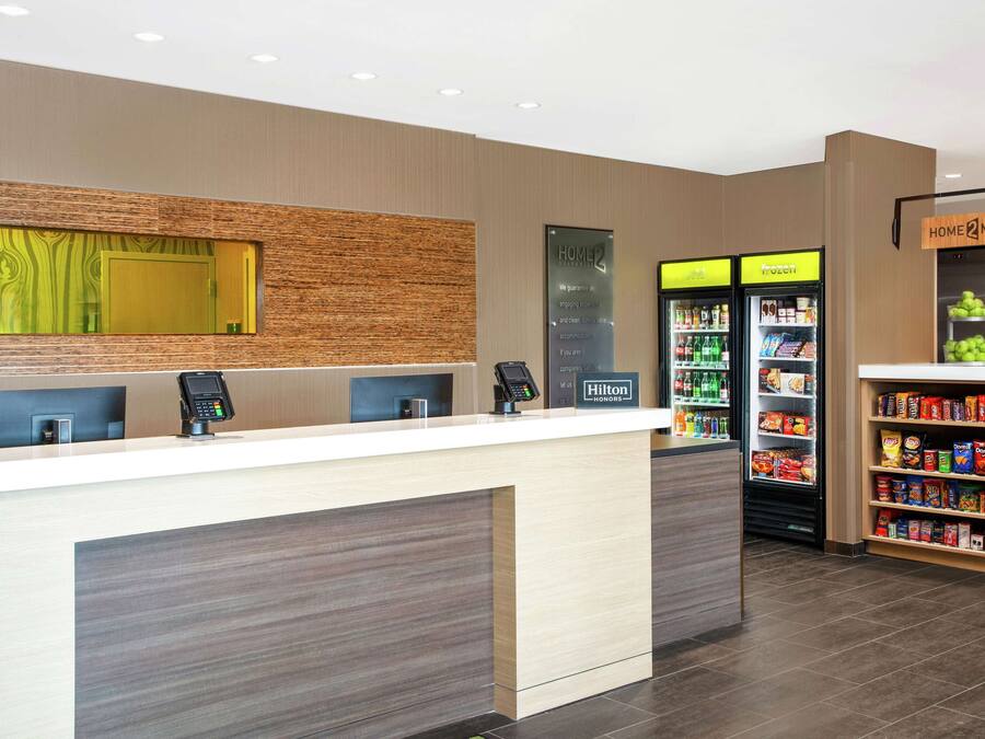Zona de recepción de un hotel con diseño moderno, que incluye un mostrador de recepción, un terminal de pago digital, una nevera y una estación de autoservicio para bebidas.