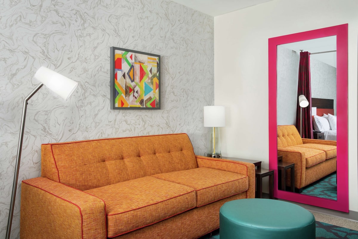 A hotel room with a sofa, a lamp, a mirror, and a colorful abstract painting.