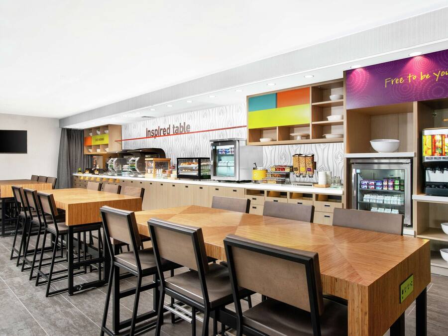 A modern cafeteria with long wooden tables and chairs, a self-service food station, and a refrigerator with beverages.
