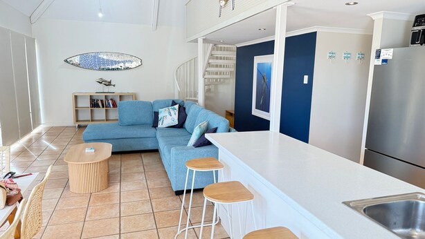 Un espacio moderno de sala con un sofá azul, una mesa de centro de madera, un taburete blanco, una cocina con refrigerador de acero inoxidable y una escalera.
