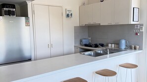 A modern kitchen with a stainless steel refrigerator, a toaster, a kettle, and a sink. There are three stools at the counter.