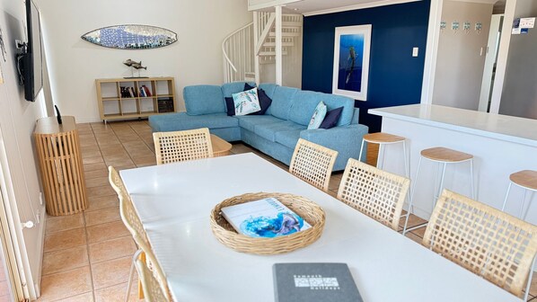 A living room with a sofa, a dining table with chairs, a bookshelf, and a staircase.