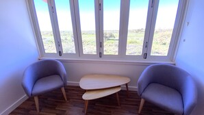 Una habitación con dos sillones grises y una pequeña mesa de madera, una ventana grande con marcos blancos, y vista a un paisaje cubierto de césped.