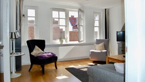 Una sala de estar con un sillón negro, un sofá gris y una mesa de centro de madera. Hay dos ventanas con cortinas, un radiador y un televisor sobre un mueble.