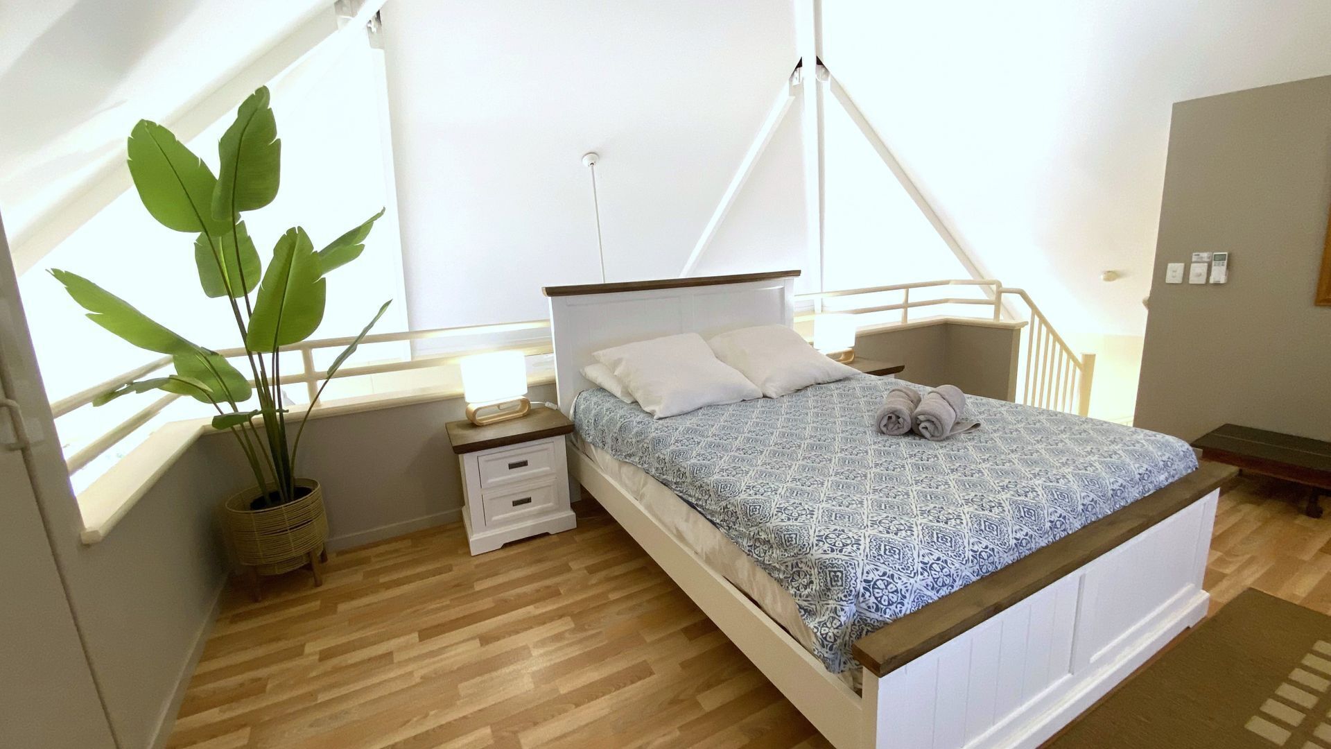 A bedroom with a bed, bedside table, nightstand, potted plant, and a window with curtains.