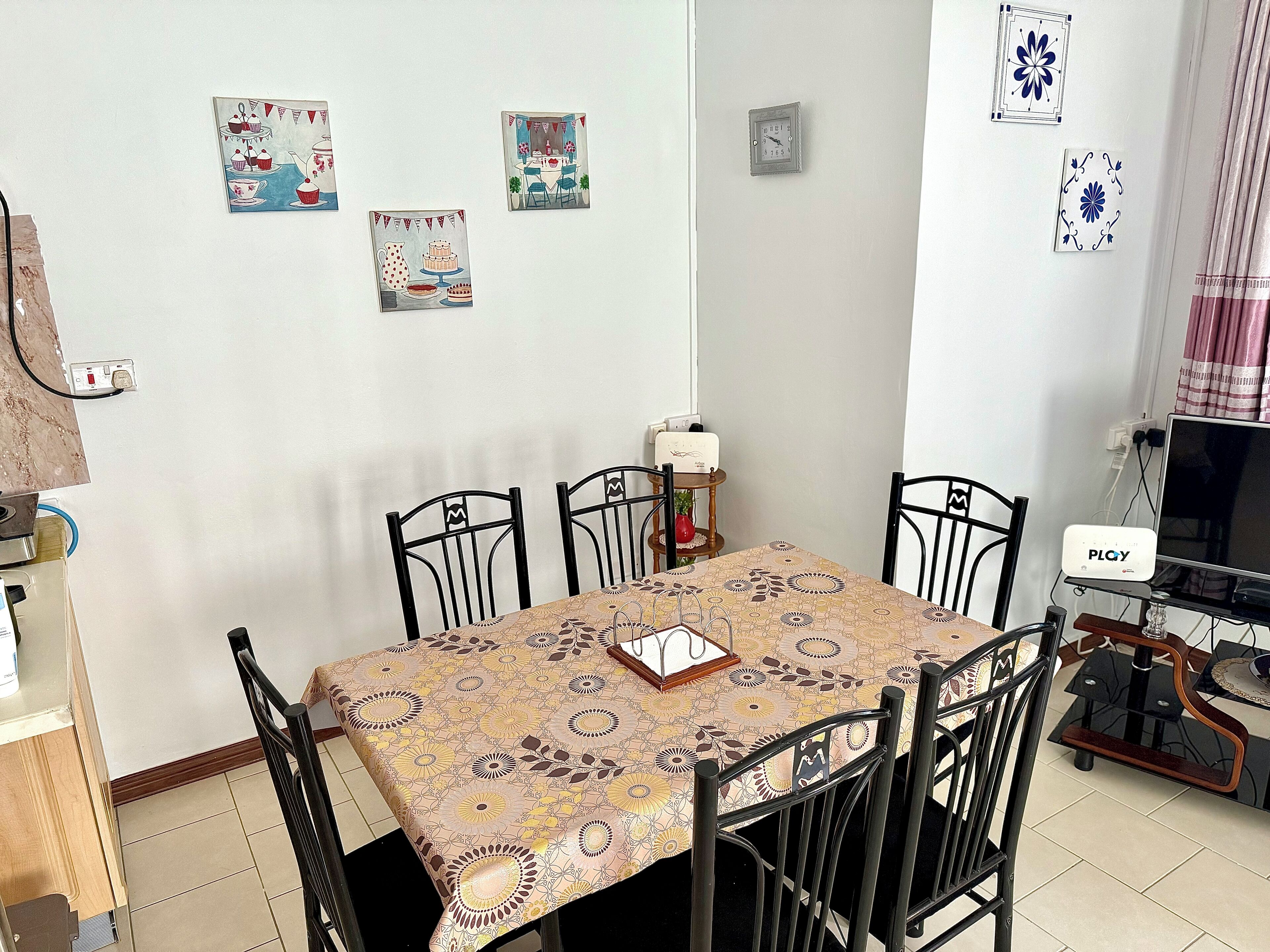 Uma área de jantar com uma mesa coberta por uma toalha estampada, rodeada por quatro cadeiras pretas. Há decorações nas paredes, um relógio e um dispositivo PLOY em um suporte.