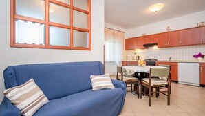 A living room with a blue sofa, a dining table with chairs, a kitchen area with wooden cabinets, and a large window with blinds. - Adriana by Interhome (Pula)