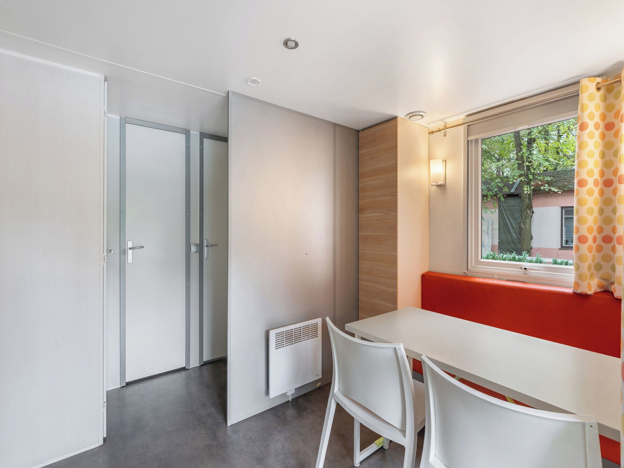 A modern room with a white table, two chairs, a bench, and a window with a view of greenery outside.