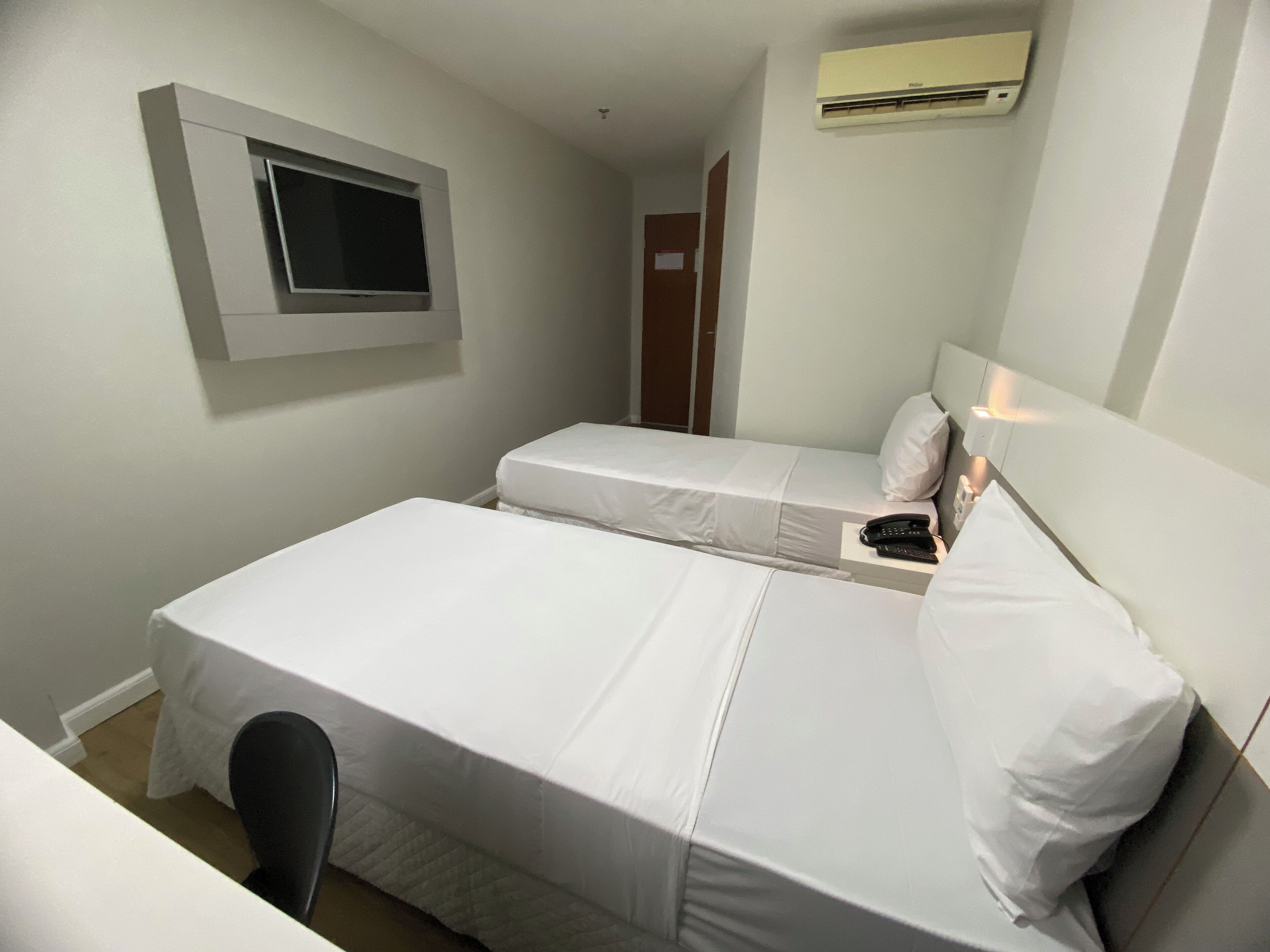 A small, clean room with two single beds, a desk, a chair, a TV mounted on the wall, and an air conditioning unit.