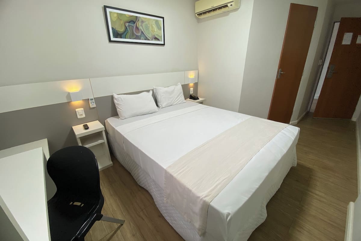 A hotel room with a bed, bedside tables, a desk, a chair, an air conditioner, and a painting on the wall.