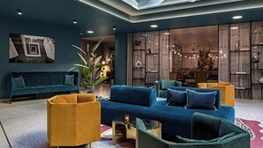 Ein modernes Wohnzimmer mit einer blauen Sofagarnitur, gelben Sessel und einem grünen Sessel. Im Hintergrund befindet sich eine große Topfpflanze und ein Essbereich.