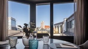 Ein Esstisch mit einer Vase, einem Glas und einem Notizbuch. Der Tisch steht in der Nähe eines Fensters mit Blick auf Gebäude und einen Turm.