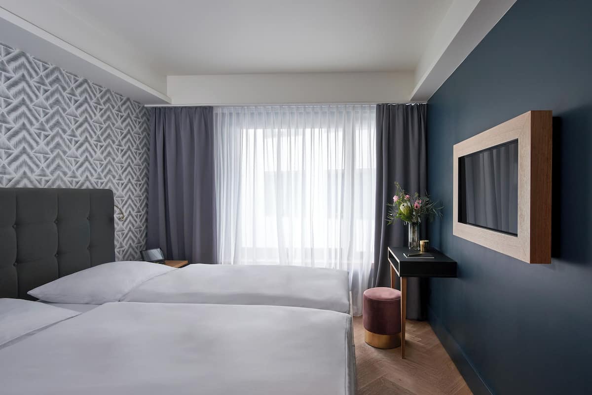 A modern hotel room with two beds, a large window with sheer curtains, a bedside table with a vase of flowers, and a wall-mounted TV.