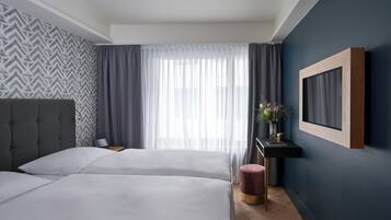 Una habitación de hotel moderna con dos camas, un ventanal con cortinas translúcidas, una mesita de noche con un jarrón de flores y un televisor montado en la pared.