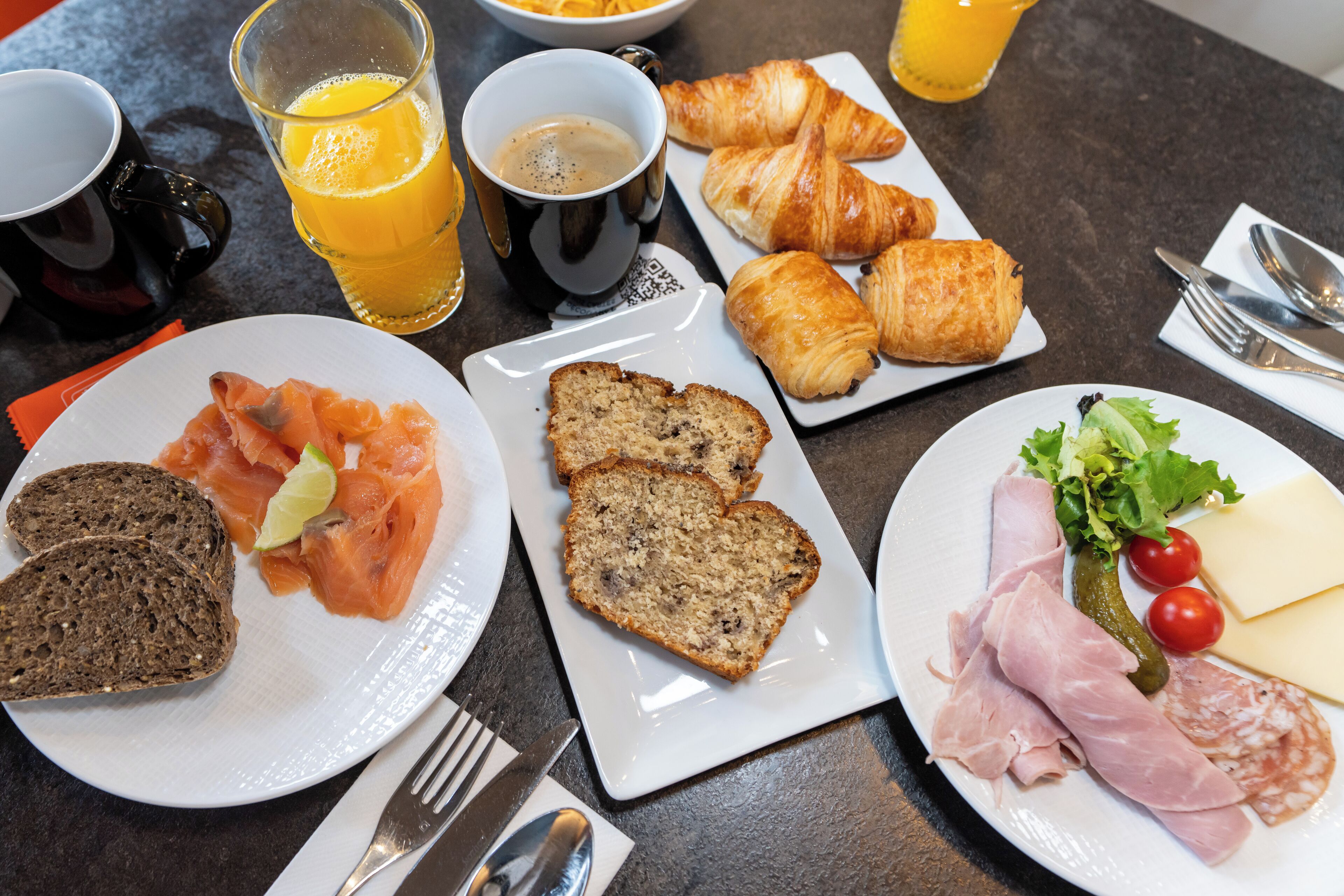 Une table dressée pour le petit-déjeuner, avec des assiettes remplies de nourriture, notamment des croissants, du saumon fumé et du pain, ainsi que des boissons et des couverts.